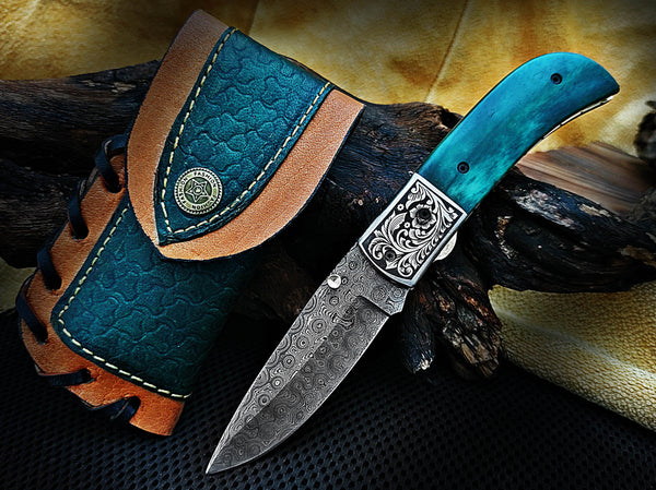 Folding knife Hand Engraved with blue and silver design, leather sheath, on wooden background
